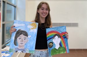 A high school aged female intern smiling while holding two pieces of elementary school student artwork.
